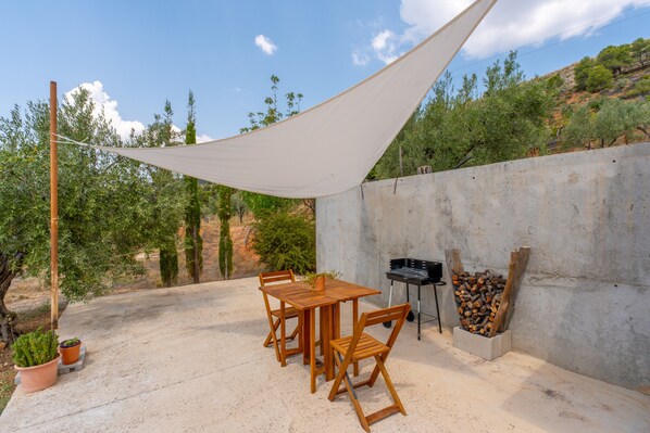 An outdoor patio with a wooden table and chairs, a barbecue grill, and a pile of firewood against a concrete wall.