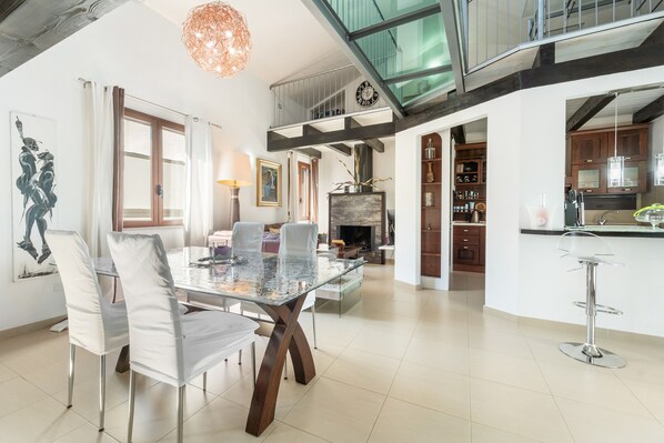 A spacious living area with a glass dining table, modern chairs, a fireplace, and a kitchen with wooden cabinets.