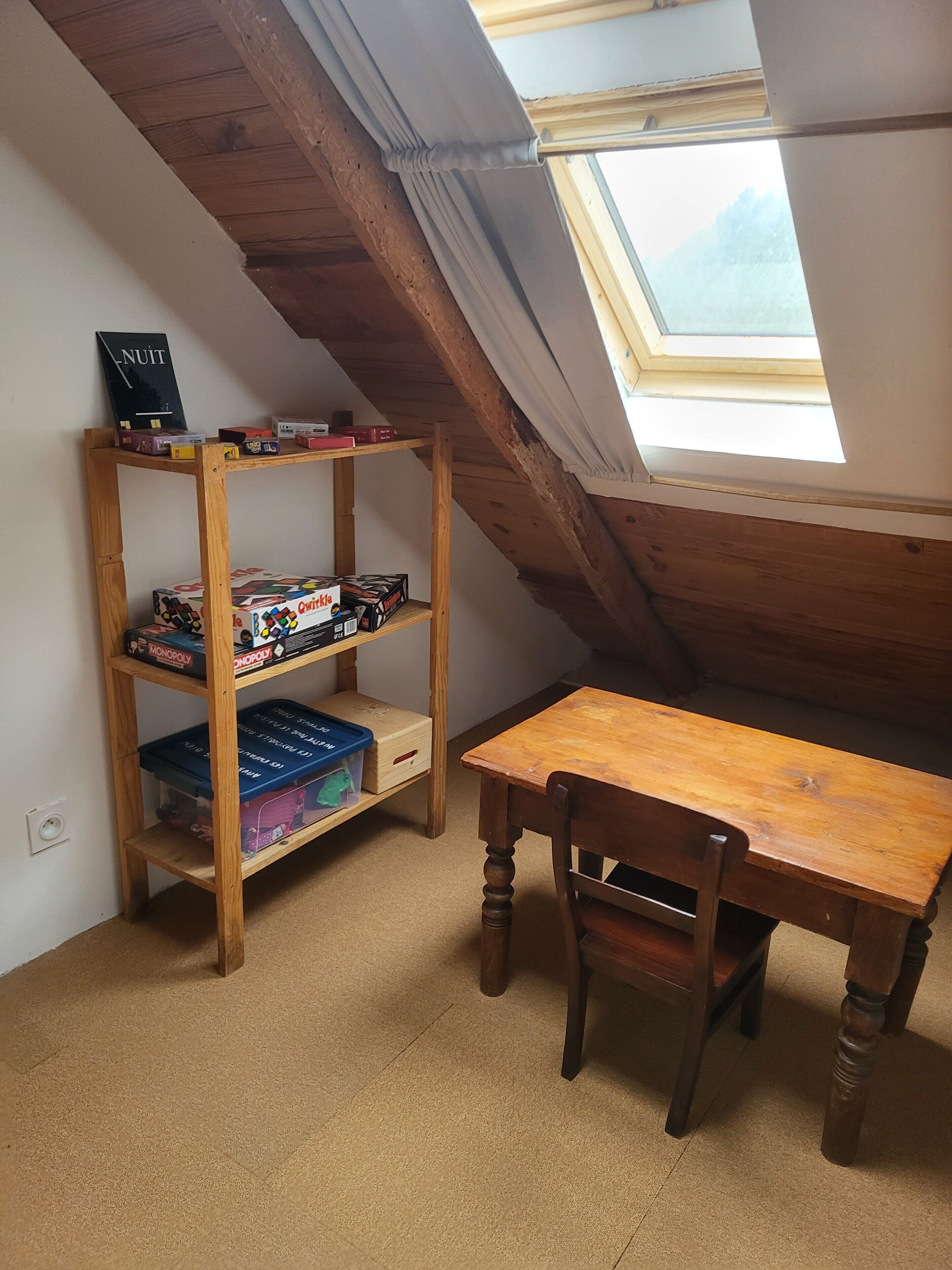 Een kamer met een schuine kap, dakraam, een houten tafel, een stoel en een boekenkast met diverse spullen.