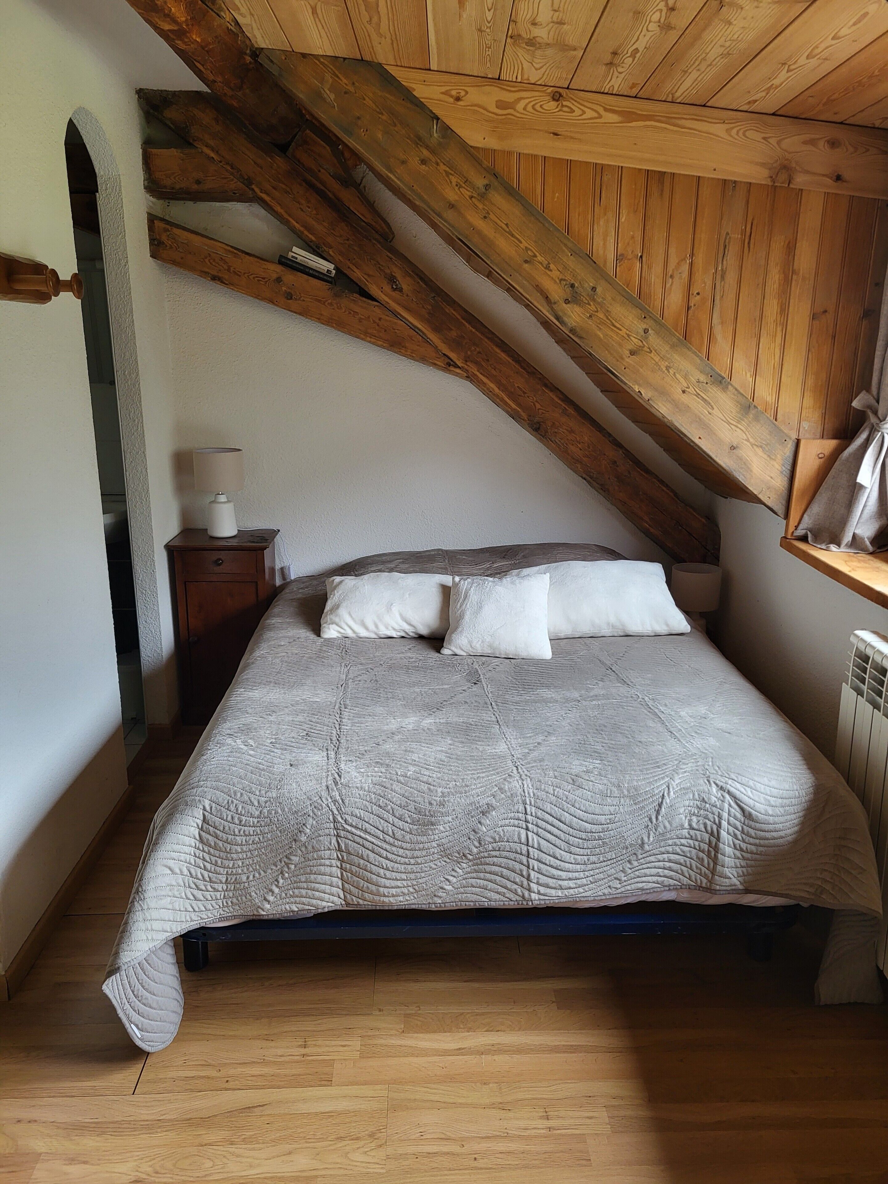 A bedroom with a sloped ceiling, a bed with a quilted blanket, two pillows, a bedside table with a lamp, and a radiator.