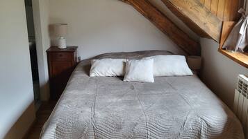 A bedroom with a sloped ceiling, a bed with a quilted blanket, two pillows, a bedside table with a lamp, and a radiator.