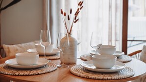 Un tavolo rotondo in legno con piatti bianchi, ciotole e bicchieri da vino, un vaso bianco con piante secche e sottopiatti intrecciati.