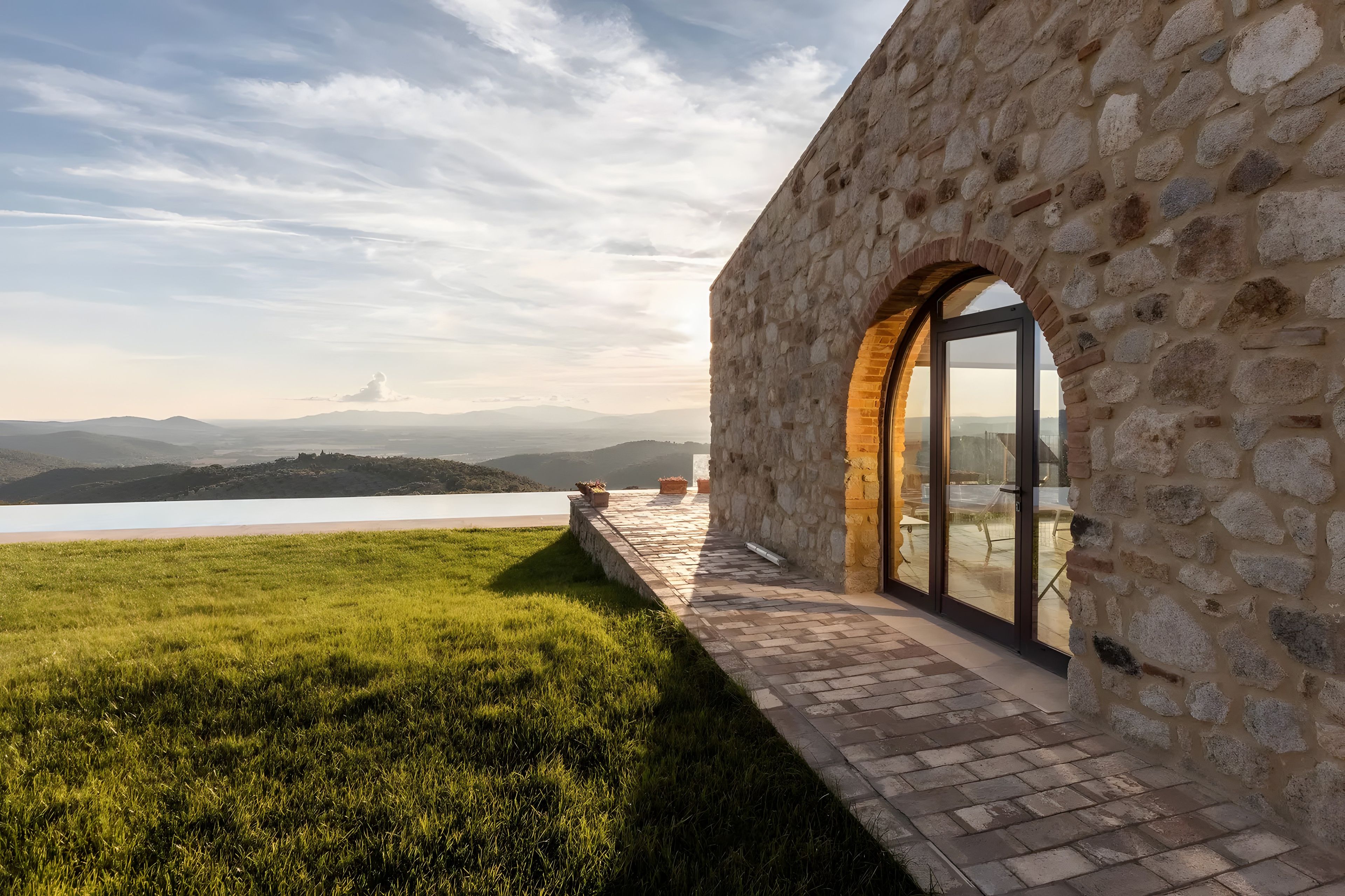 A stone building with a large arched window overlooking a grassy area and a body of water.