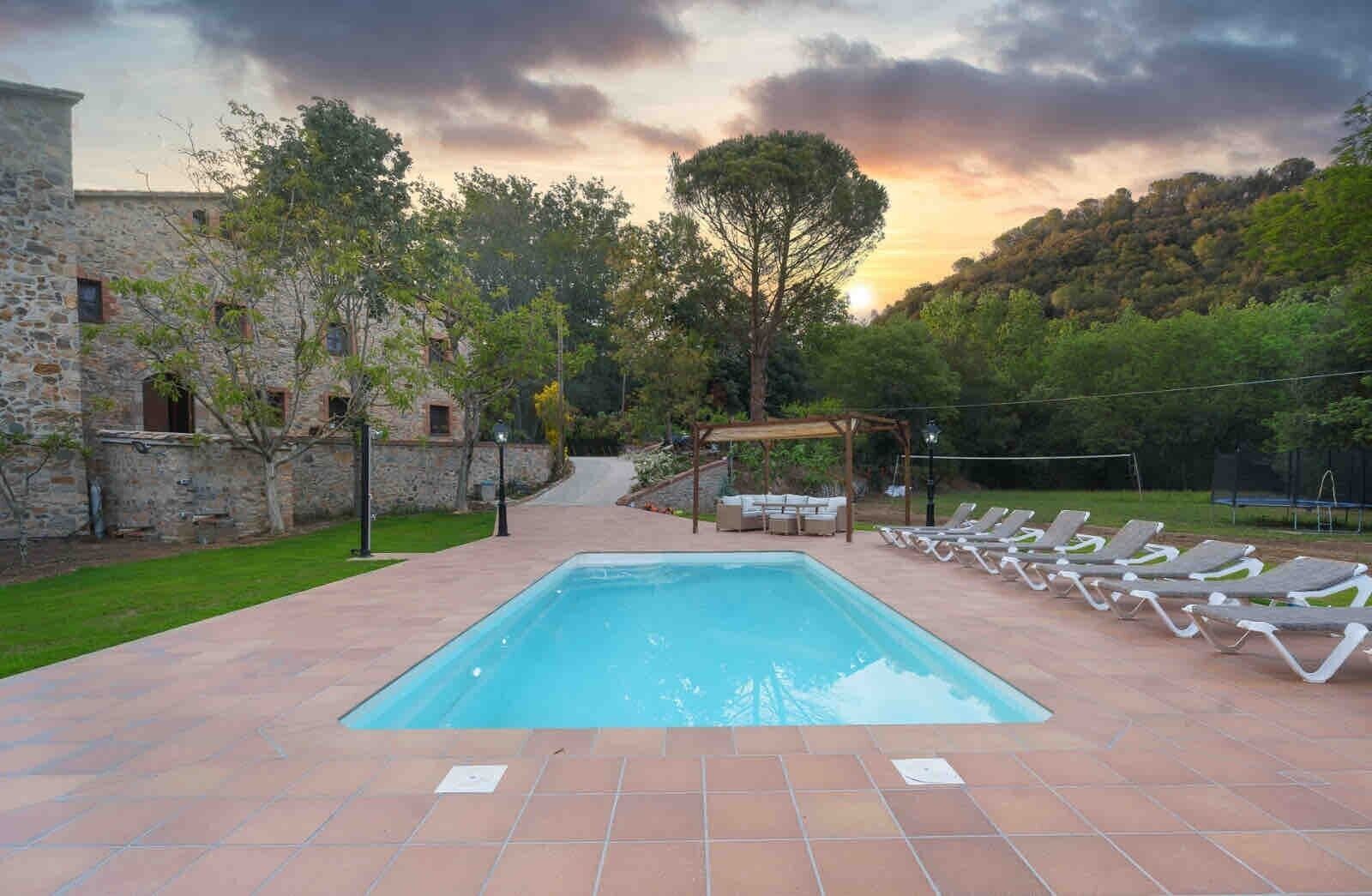 Una piscina rectangular con una pérgola y sillones de descanso al fondo.