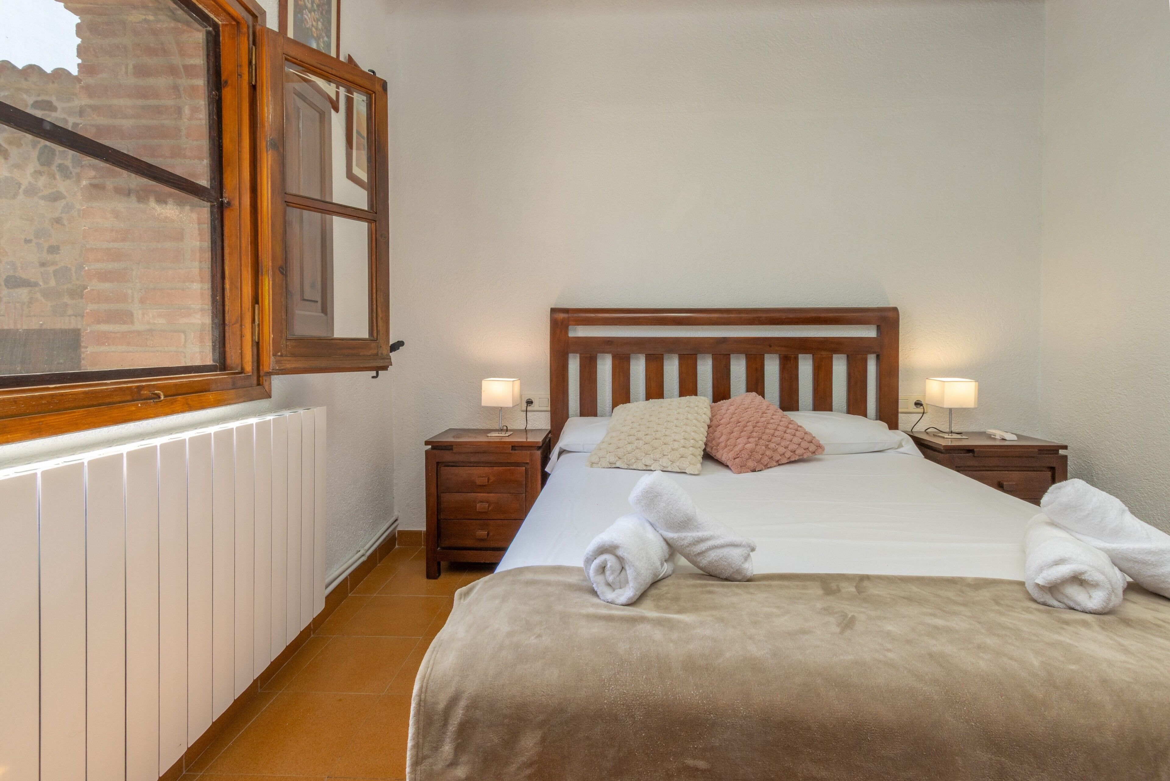Ein Schlafzimmer mit einem hölzernen Bett, zwei Nachttischen mit Lampen, einem Heizkörper und einem Fenster mit Holzjalousien.