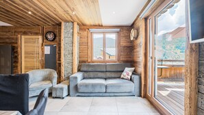 A cozy room with a grey sofa, a small table, and a chair. There is a wooden ceiling, a stone wall, and a large window. - Apartment "Le Galibier" with Mountain View, Balcony and Wi-Fi (Valloire)