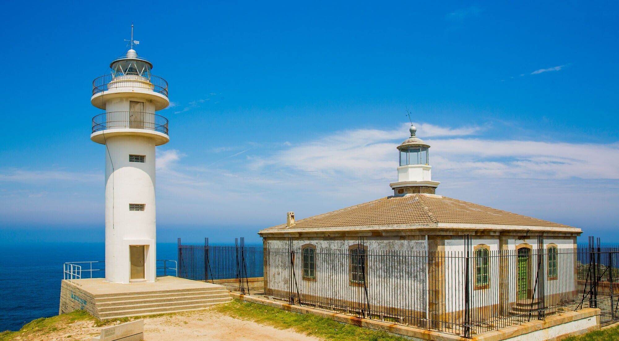 Un faro y un edificio con tejado de baldosas, ambos con barandillas de metal, junto al mar.