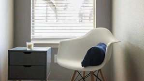 A room with a window, a white chair with a blue cushion, and a small table with a glass of milk.