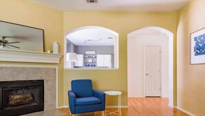 A living room with a fireplace, a blue armchair, a small table, and a kitchen visible through an archway.