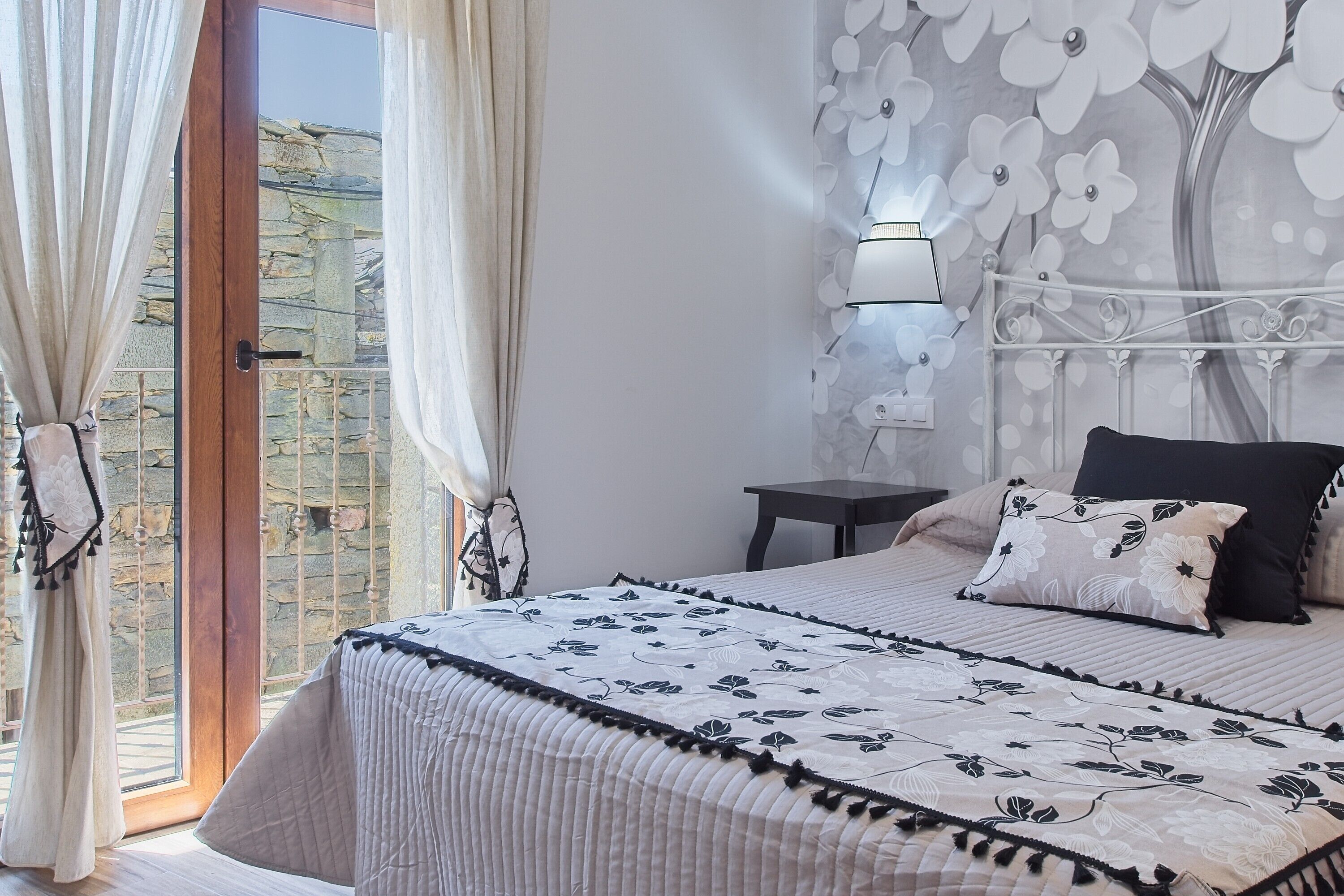 A bedroom with a bed, a nightstand, a wall-mounted lamp, and a view of a stone wall through a glass door.