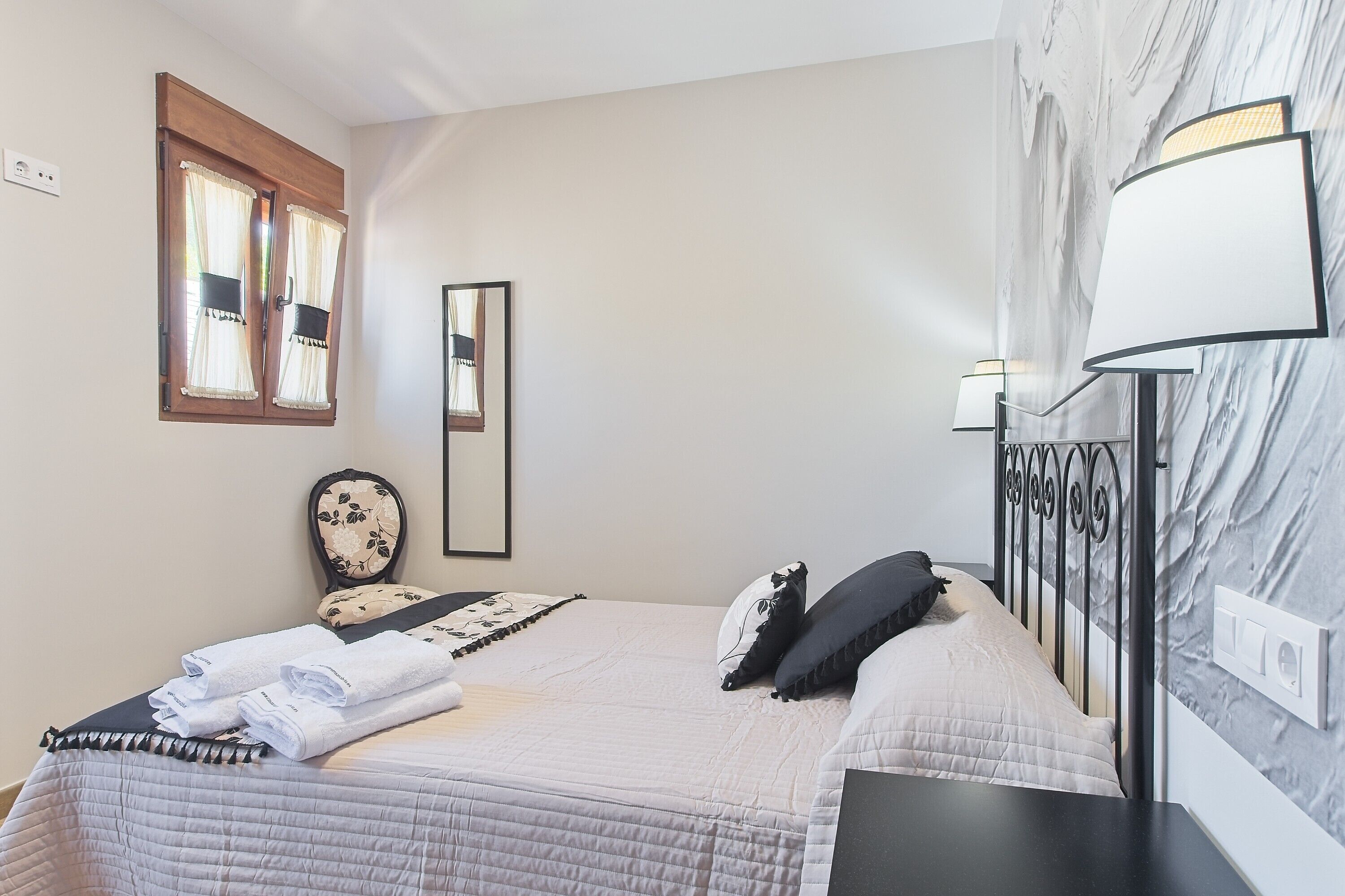 Un lit bien fait, recouvert d’une courtepointe, une table de chevet avec une lampe, un miroir et une fenêtre avec des rideaux.