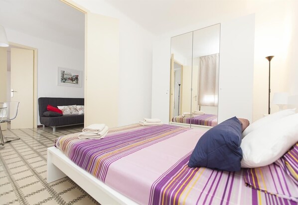 A hotel room with a bed, a chair, a desk, a mirror, and a painting on the wall.