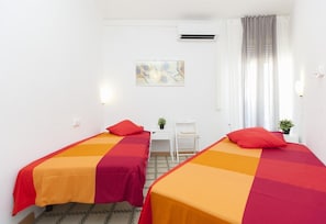 A hotel room with two beds, a desk, a chair, a window with curtains, and an air conditioning unit.