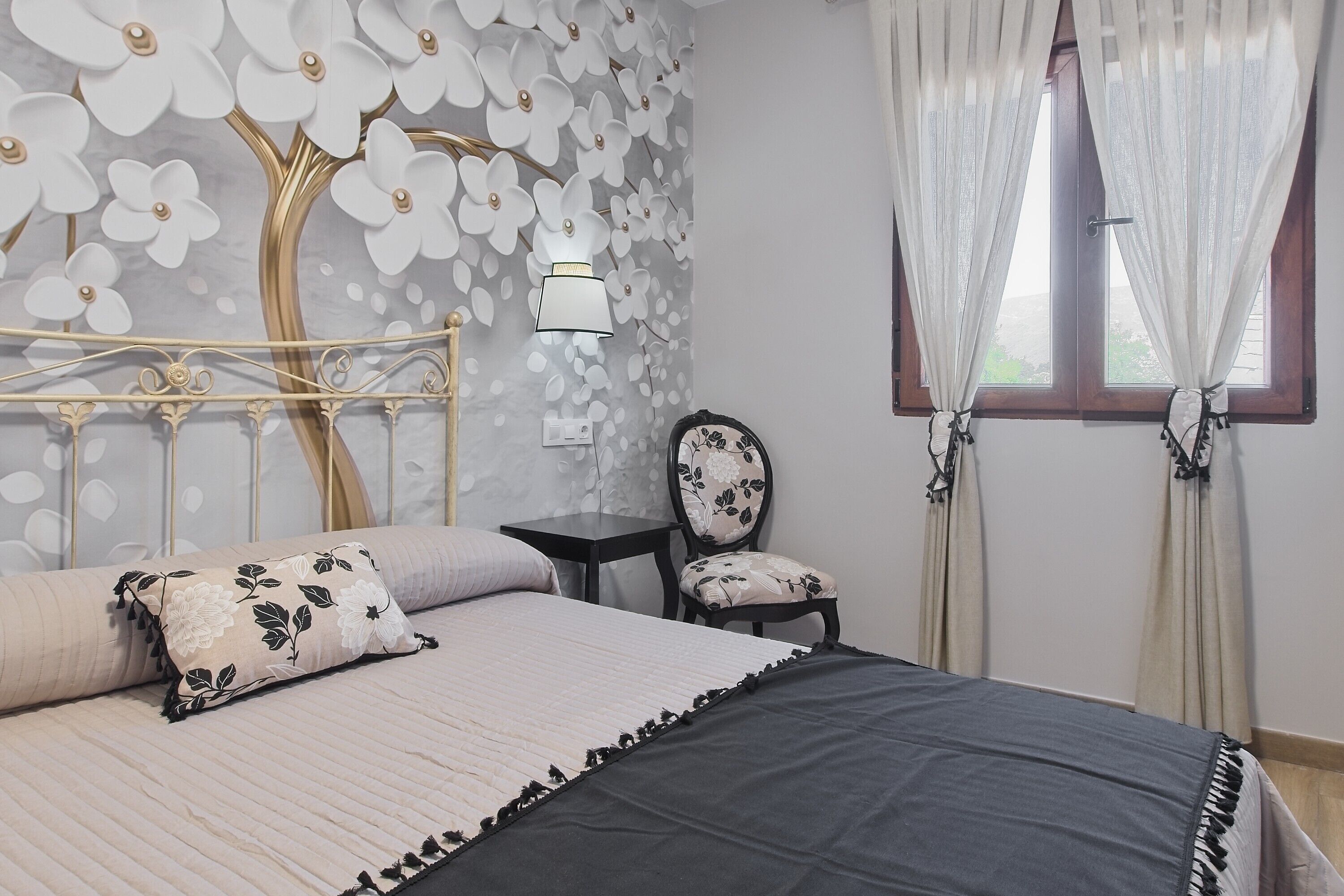 Ein Schlafzimmer mit Bett, Stuhl, Nachttisch, Wand mit Blumenmuster und Fenster mit Vorhängen.