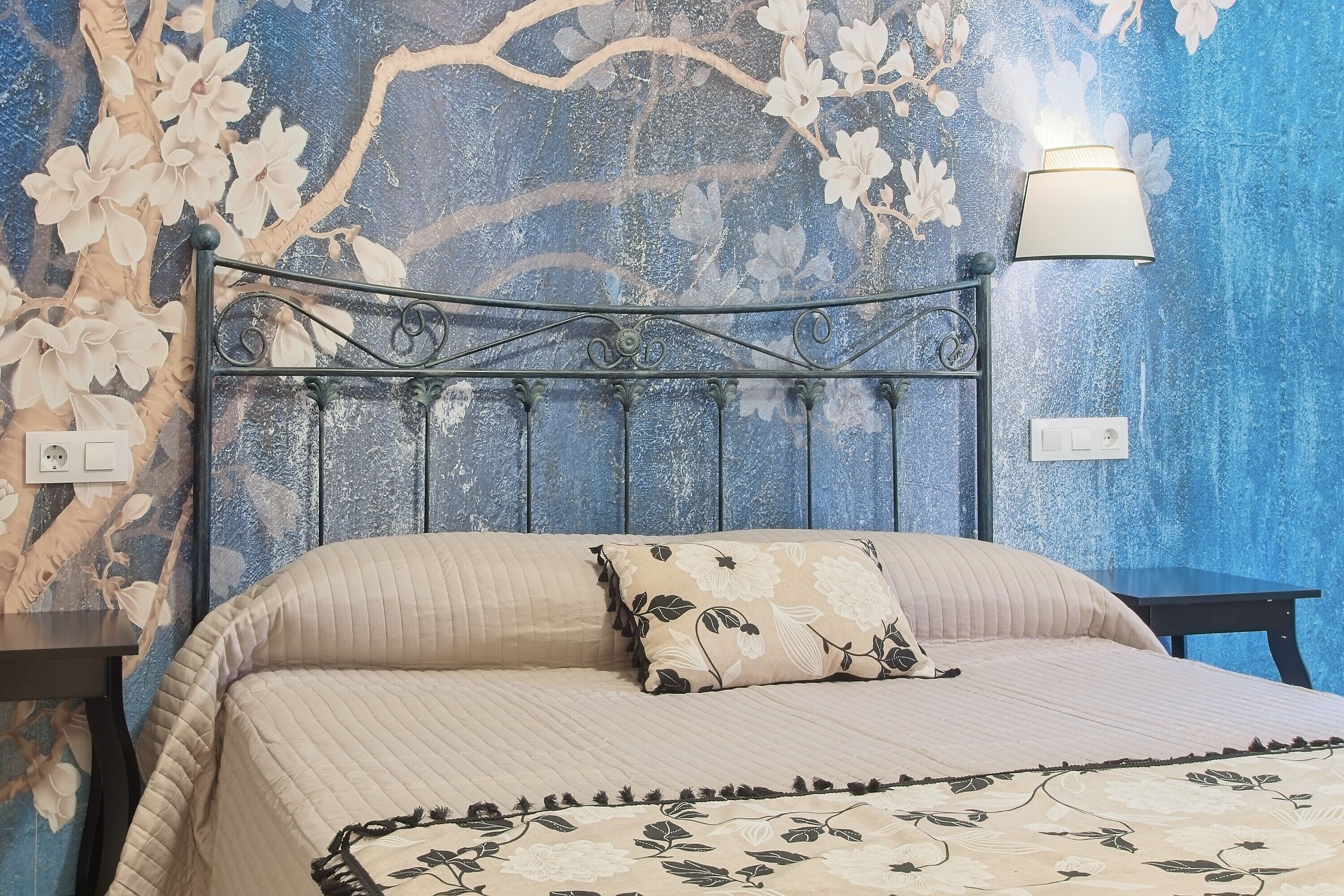 A bedroom with a metal headboard, floral wallpaper, a bedside lamp, and a bed with a floral bedspread.