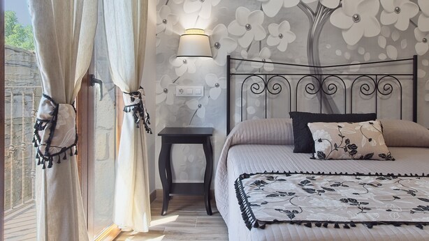 A bedroom with a bed, curtains, a nightstand, a lamp, and a wall with a floral pattern. - Holiday Home 'Lago De Sanabria' with Mountain View, Private Garden and Balcony (Coso)