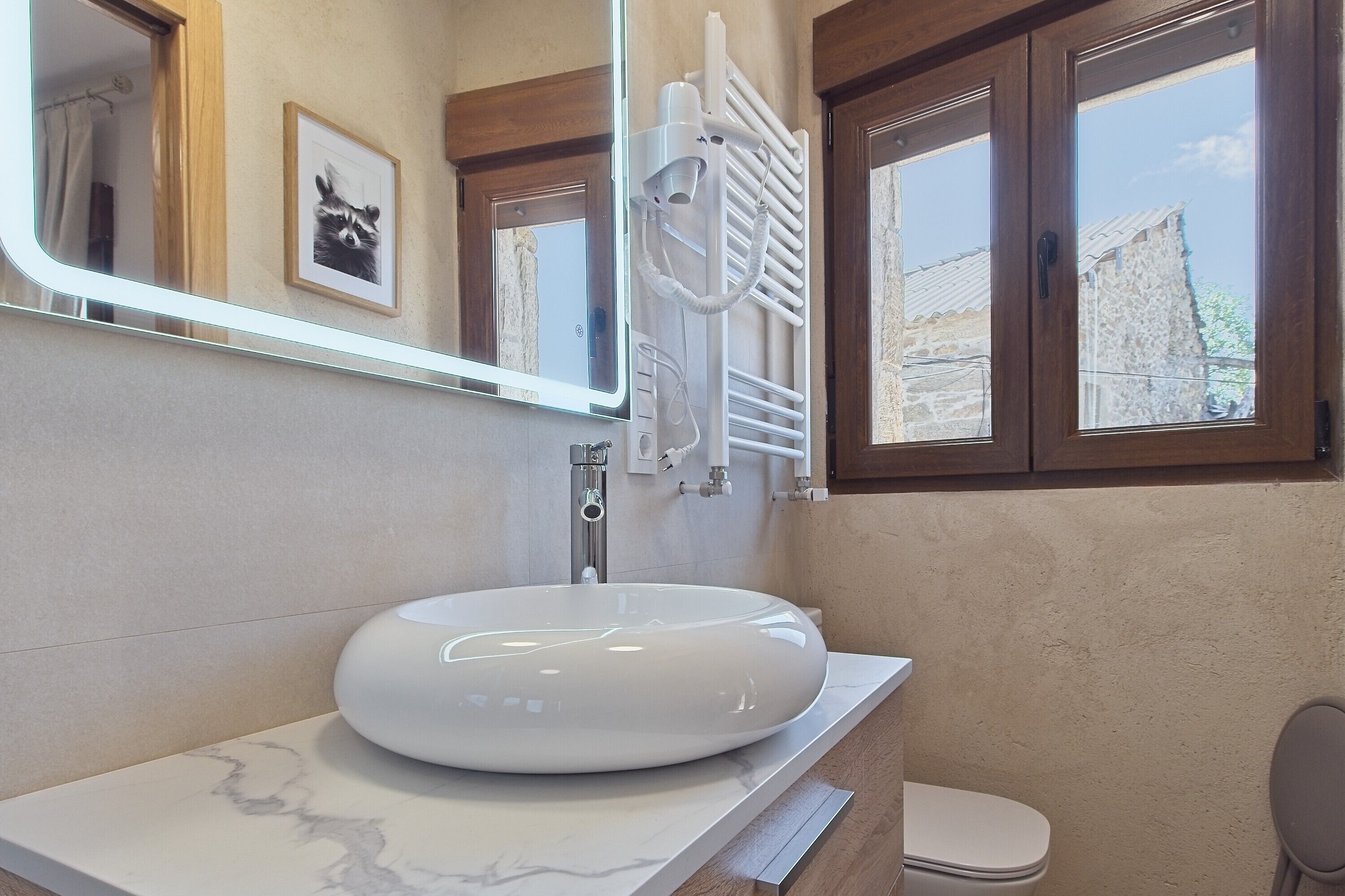 Une salle de bain avec un lavabo blanc, un miroir, un porte-serviettes et une fenêtre donnant sur un immeuble.