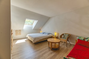 A room with a bed, a round wooden table, a wicker chair, and a small window.