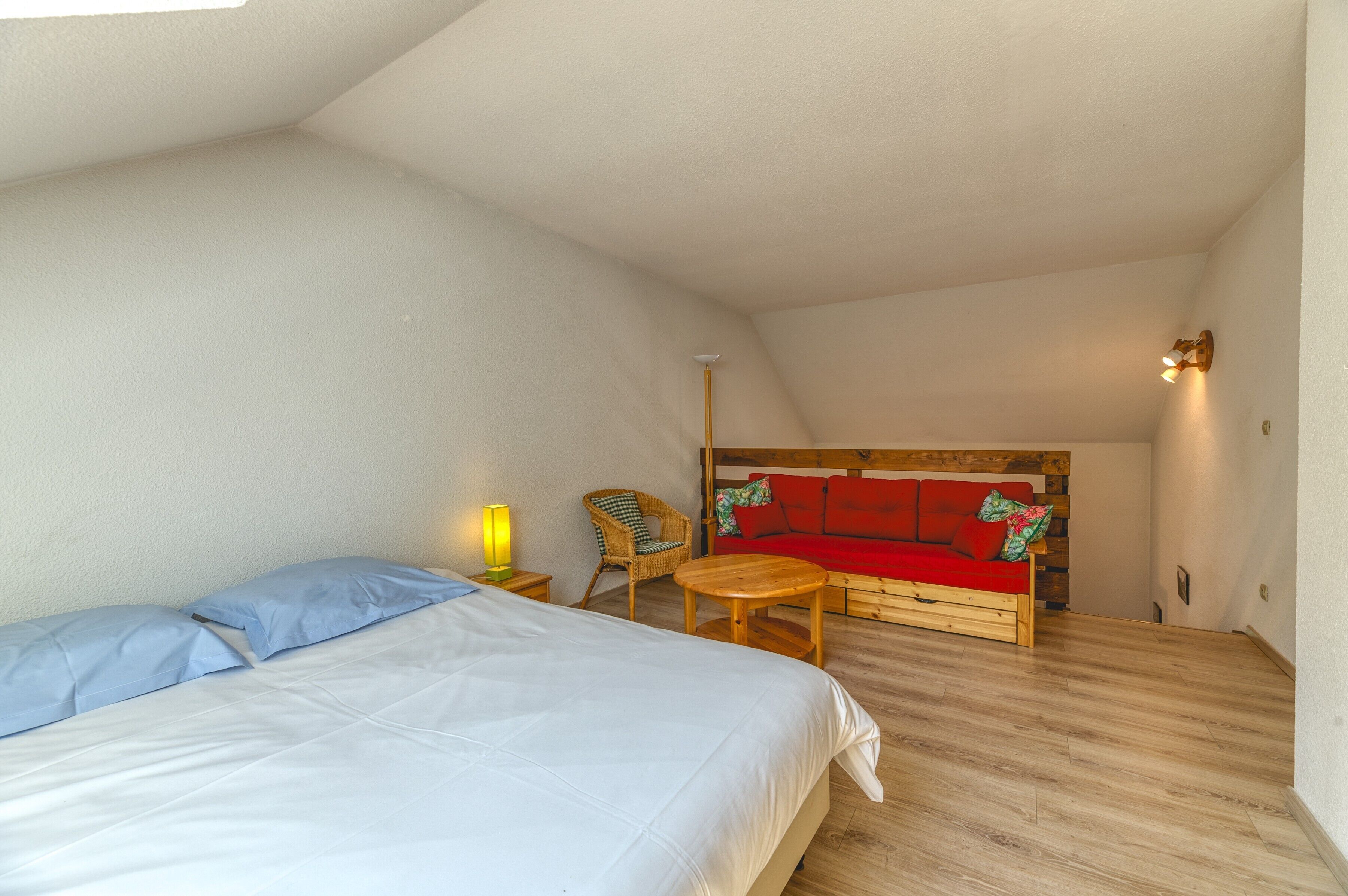 Une chambre à coucher avec un lit, un canapé rouge, une table en bois, une chaise en osier et une lampe.