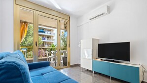 A living room with a blue sofa, a television on a teal cabinet, and a sliding glass door leading to an outdoor balcony.