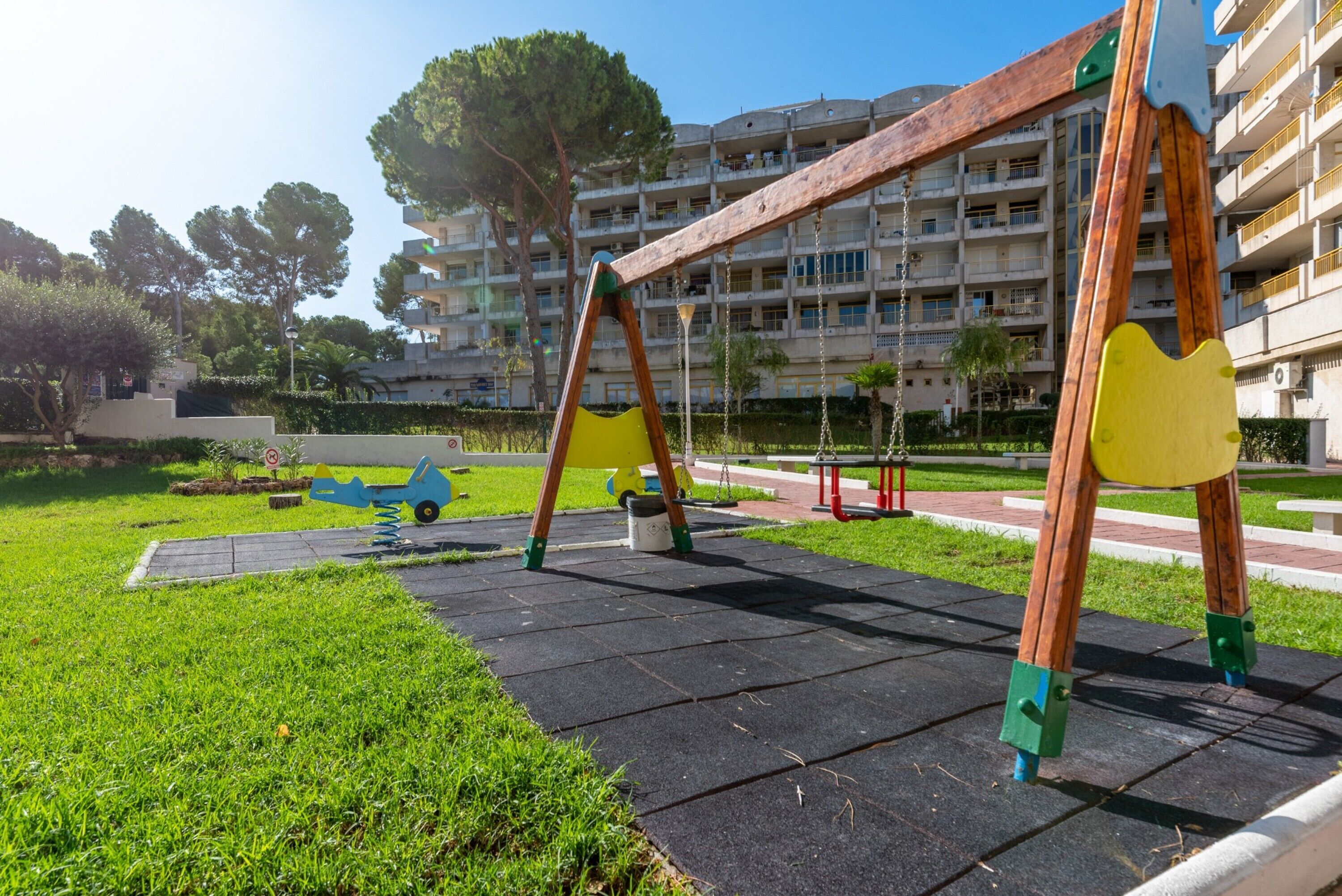 A playground with swings and a slide, surrounded by grass and a paved area, with a multi-story building in the background.