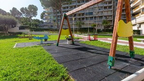 A playground with swings and a slide, surrounded by grass and a paved area, with a multi-story building in the background.