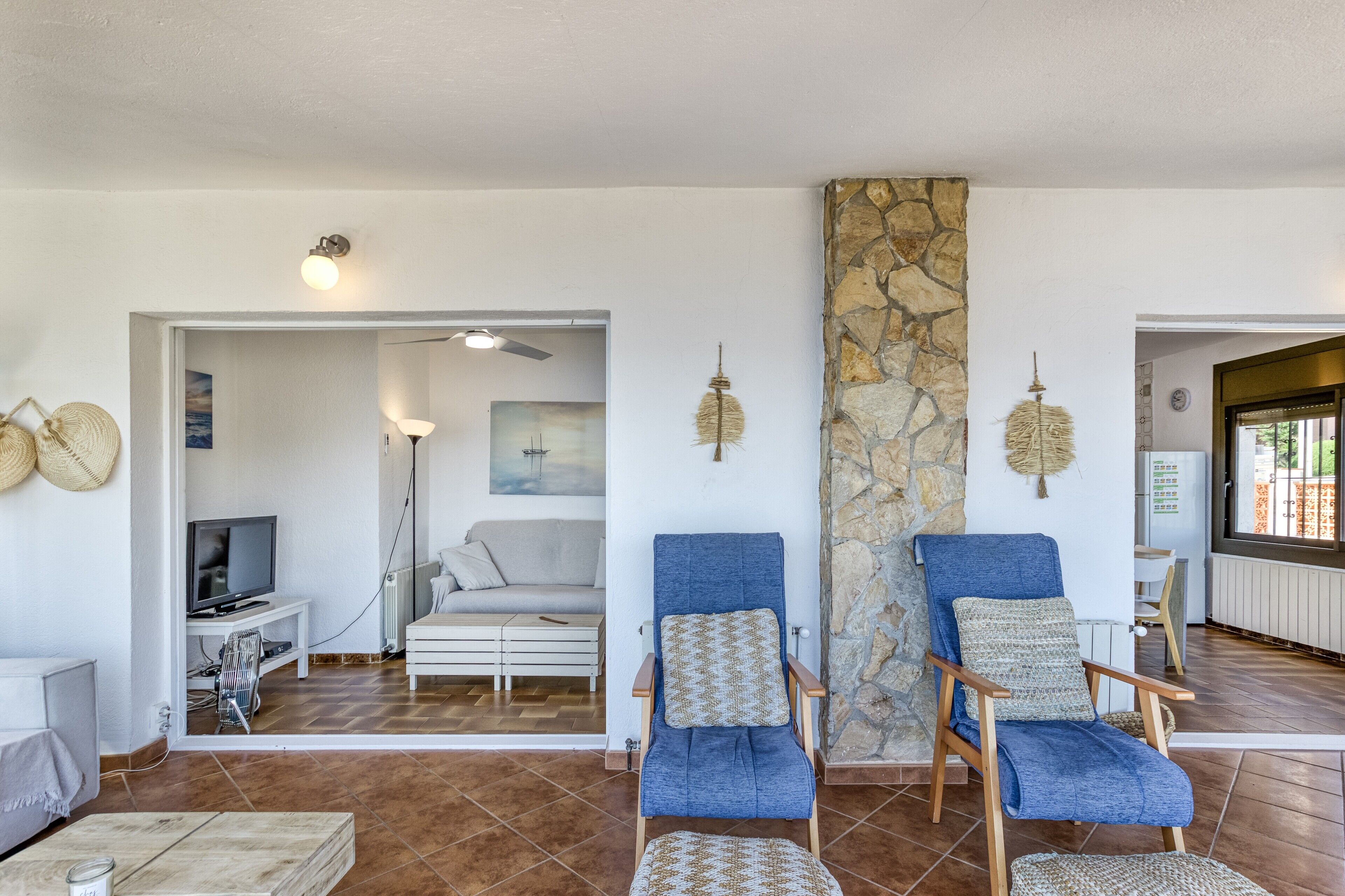 Una sala de estar con una pared de piedra, dos sillones azules, un sofá blanco, un televisor y un ventilador de techo.