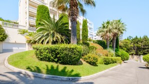 A well-maintained garden with palm trees and hedges, a curved driveway, and a multi-story building with balconies.