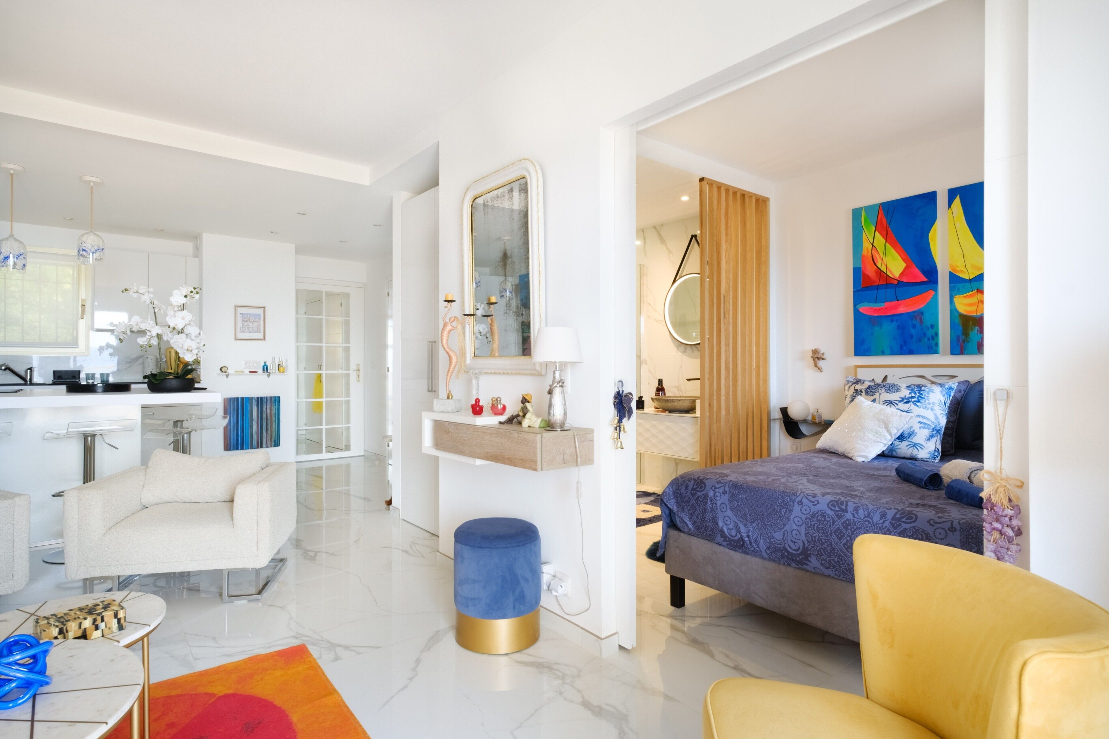 Une chambre d’hôtel moderne comprenant un lit, un coin salon avec un fauteuil, une petite table, une coiffeuse avec un miroir et des œuvres d’art accrochées aux murs.