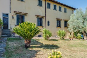 Un edificio con fachada color beige, varias ventanas con contraventanas, una palmera en primer plano y un césped bien cuidado.