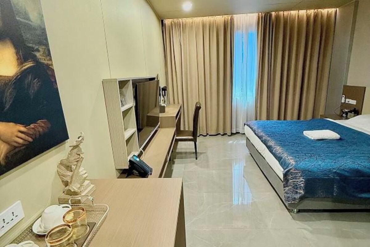 A hotel room with a bed, a TV, a desk with a tray of glasses, and a painting on the wall.