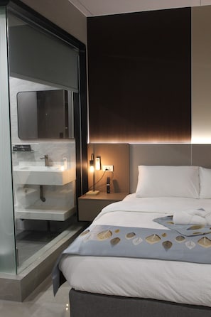 A modern hotel room with a glass-enclosed shower, a bedside table with a lamp, and a bed with a patterned bedspread. - Skyport Hotel (Pasay)