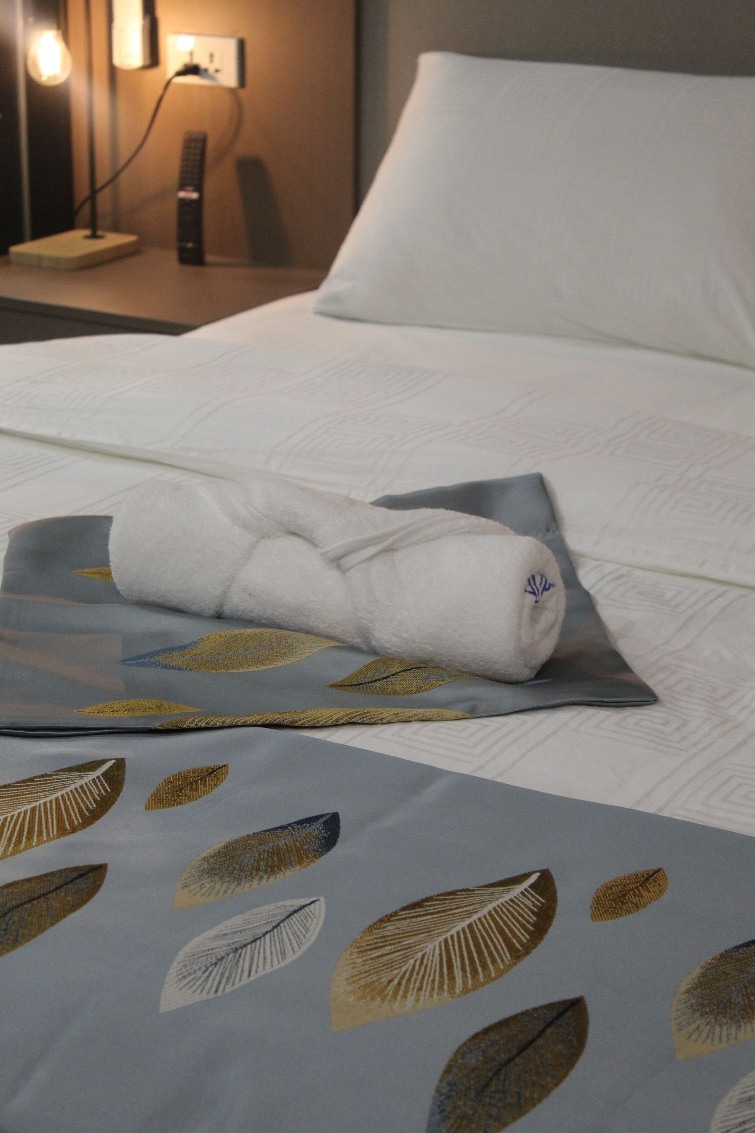 A neatly made bed with a rolled white towel and a pillow on a bed with a leaf-patterned bedspread.
