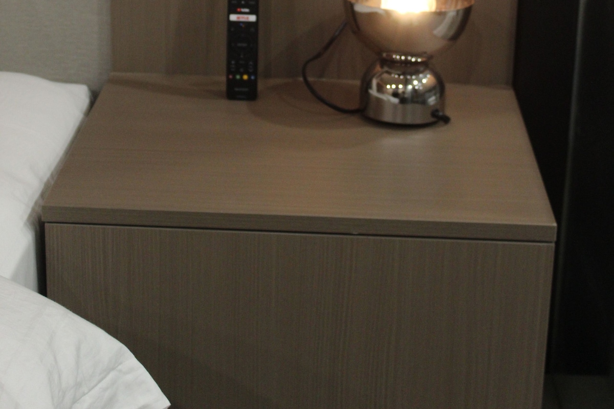 Habitación de hotel con mesita de noche, lámpara, control remoto y estación de carga en la pared.