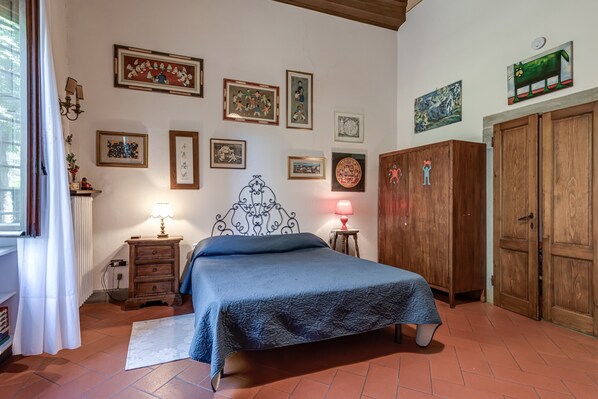 A bedroom with a bed, wall-mounted artwork, a wooden wardrobe, and a nightstand with a lamp.