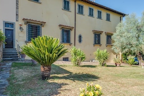 Un bâtiment à la façade beige, doté de nombreuses fenêtres avec volets, un palmier au premier plan et une pelouse bien entretenue.
