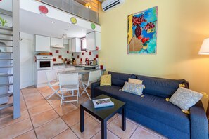 Eine moderne Küche mit weißen Schränken, ein Essbereich mit weißem Tisch und Stühlen, ein dunkelblaues Sofa mit gemusterten Kissen und ein farbenfrohes abstraktes Bild an der Wand.