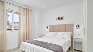 A bedroom with a bed, curtains, a nightstand, a wall-mounted lamp, and a view of buildings outside.