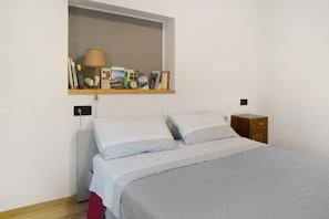 En pænt redt seng med lyseblå dyne og puder, et natbord med lampe, og en hylde over sengen med bøger og et ur.
