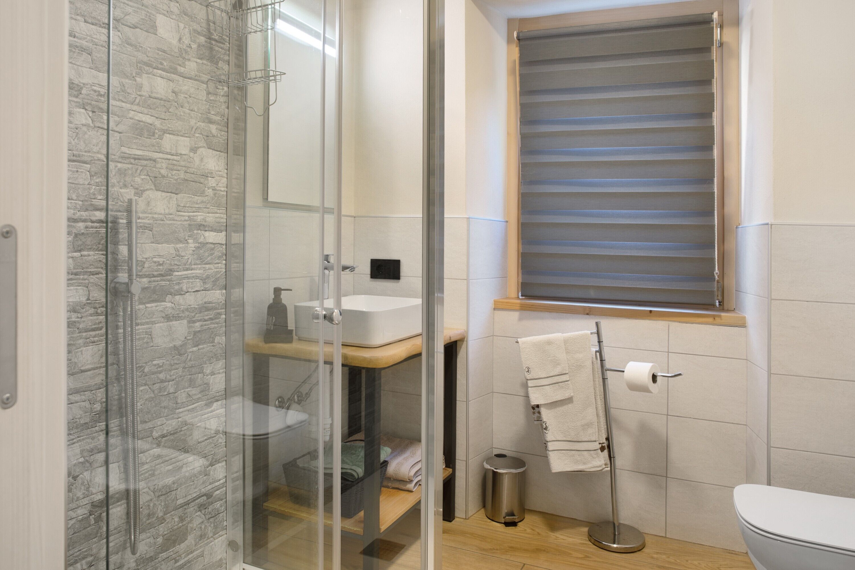 A bathroom with a glass shower enclosure, a sink, a toilet, and a window with blinds.