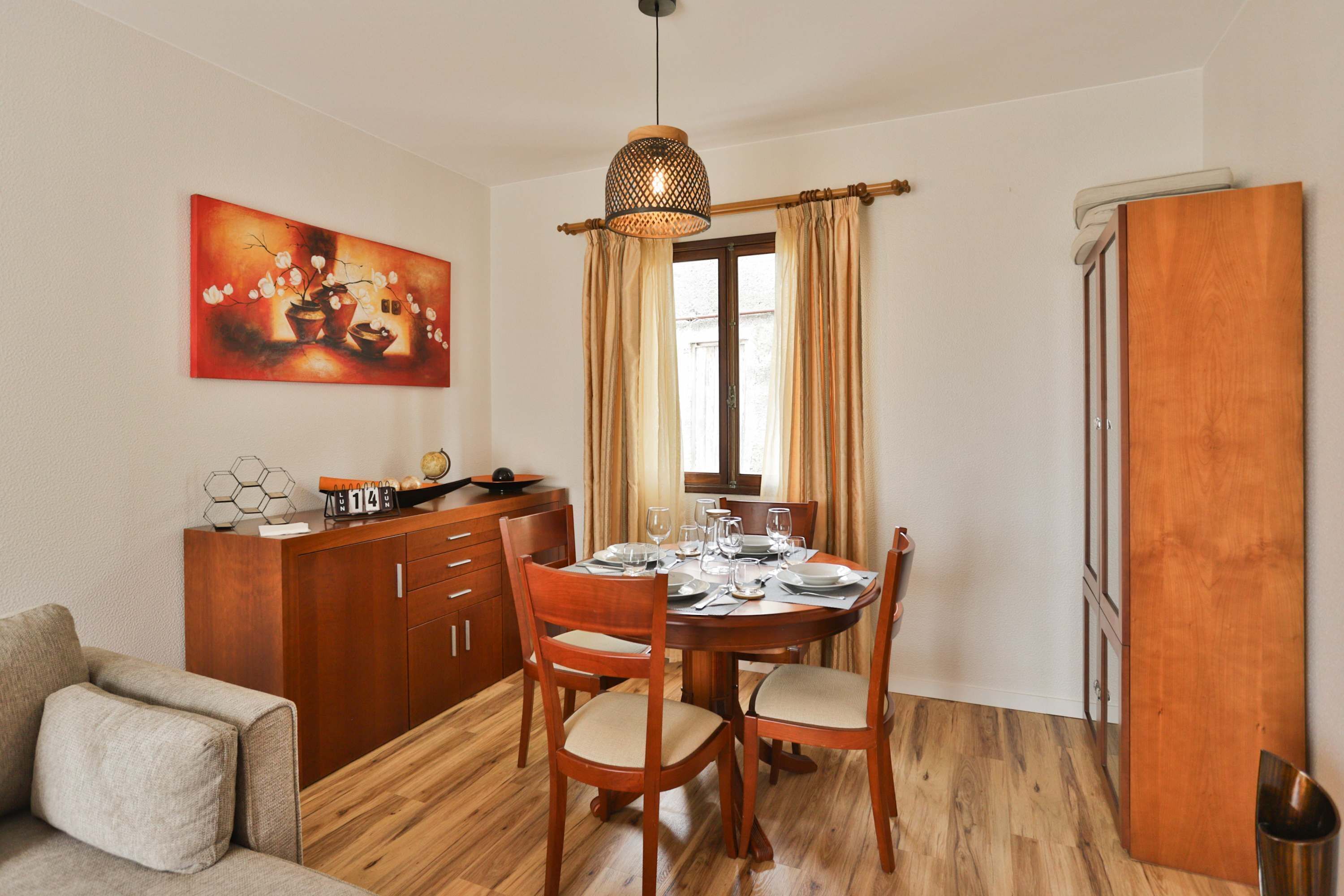 A dining area with a wooden table set for four, a wooden cabinet, a sofa, and a painting on the wall.