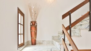 Una escalera con escalones de mármol y barandilla de madera, que lleva a un rellano con un jarrón de cobre grande y una ventana.