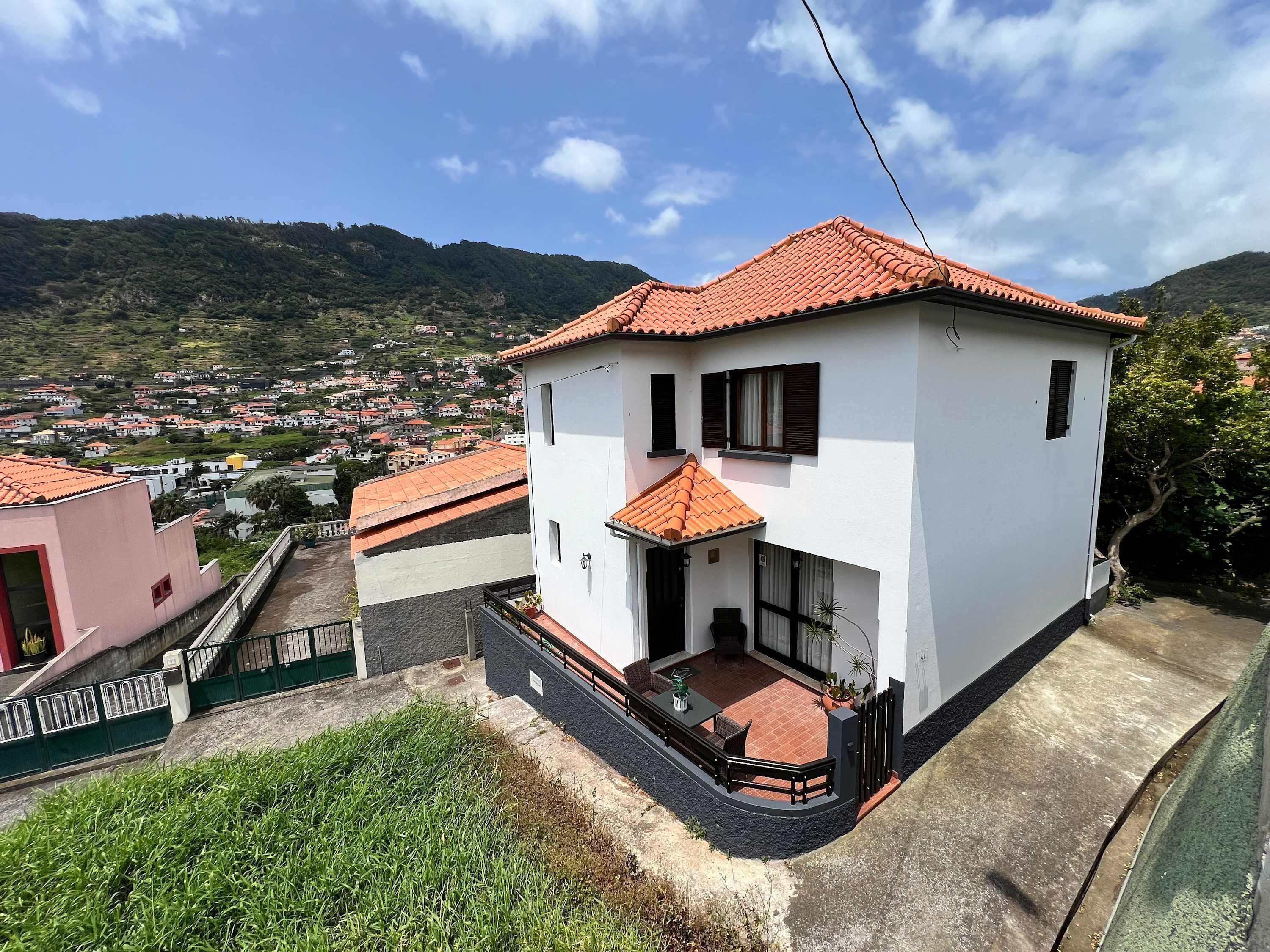 A two-story house with a red-tiled roof, a balcony, and a view of a hillside town.