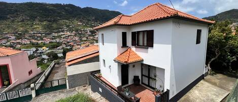 A two-story house with a red-tiled roof, a balcony, and a view of a hillside town.