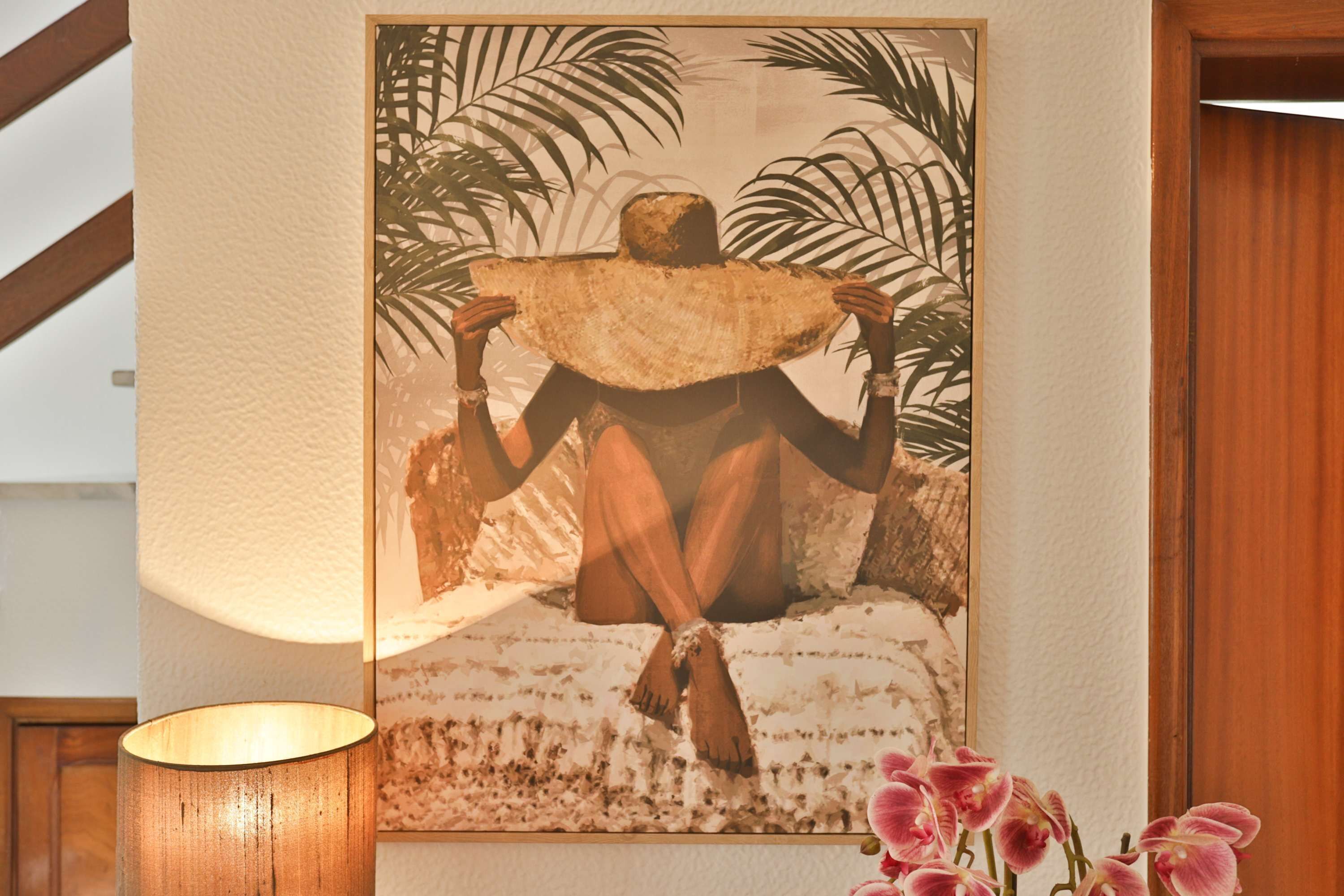 A framed artwork of a person holding a large hat, with a lamp and pink orchids in the foreground.