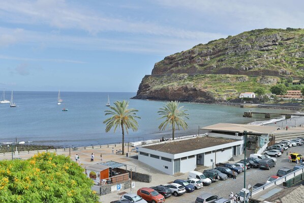 A coastal parking area with cars, a building with a flat roof, and a view of the sea with boats. - The Panoramic View in Machico (Machico)