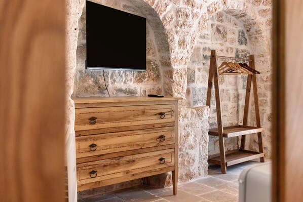 A stone-walled room with a wooden dresser, a TV mounted on the wall, and a wooden hanger stand.