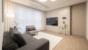 Un salon moderne avec un canapé gris, un petit fauteuil, un téléviseur à écran plat fixé au mur, une table d’appoint en verre sur laquelle est posée une plante, et une porte en bois.