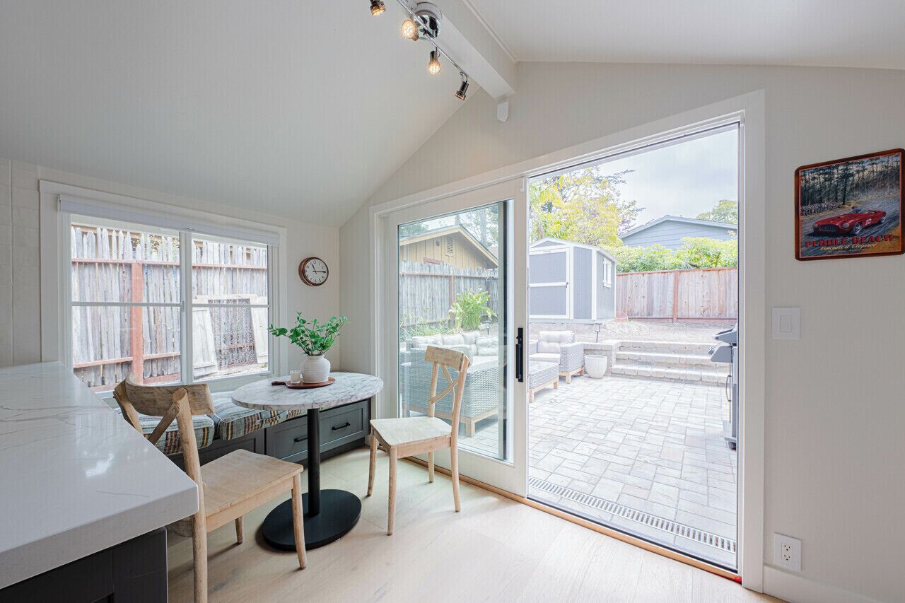 Une cuisine lumineuse avec un comptoir en marbre, des chaises en bois, une plante en pot et une porte-fenêtre coulissante donnant sur un patio aménagé avec du mobilier d’extérieur.