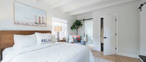 A bedroom with a large bed, a nightstand, a lamp, a chair, and a door leading to another room.